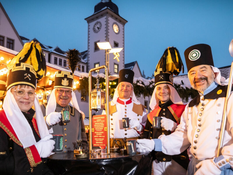 Vor stimmungsvoll beleuchteten Weihnachtsmarktständen stehen fünf Bergleute - vier Männer und eine Frau - im traditionell bergmännischen Habit mit Glühweintassen an einem Tisch. Es ist Abend. Im Hintergrund ist ein Kirchturm zu erkennen.