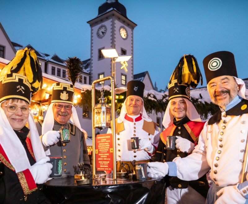 Vor stimmungsvoll beleuchteten Weihnachtsmarktständen stehen fünf Bergleute - vier Männer und eine Frau - im traditionell bergmännischen Habit mit Glühweintassen an einem Tisch. Es ist Abend. Im Hintergrund ist ein Kirchturm zu erkennen.