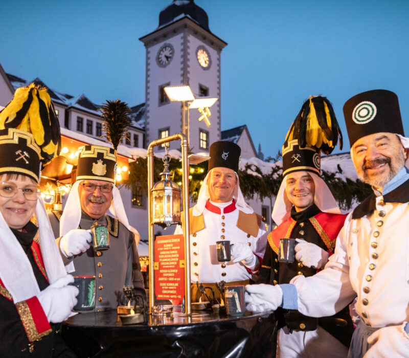 Vor stimmungsvoll beleuchteten Weihnachtsmarktständen stehen fünf Bergleute - vier Männer und eine Frau - im traditionell bergmännischen Habit mit Glühweintassen an einem Tisch. Es ist Abend. Im Hintergrund ist ein Kirchturm zu erkennen.