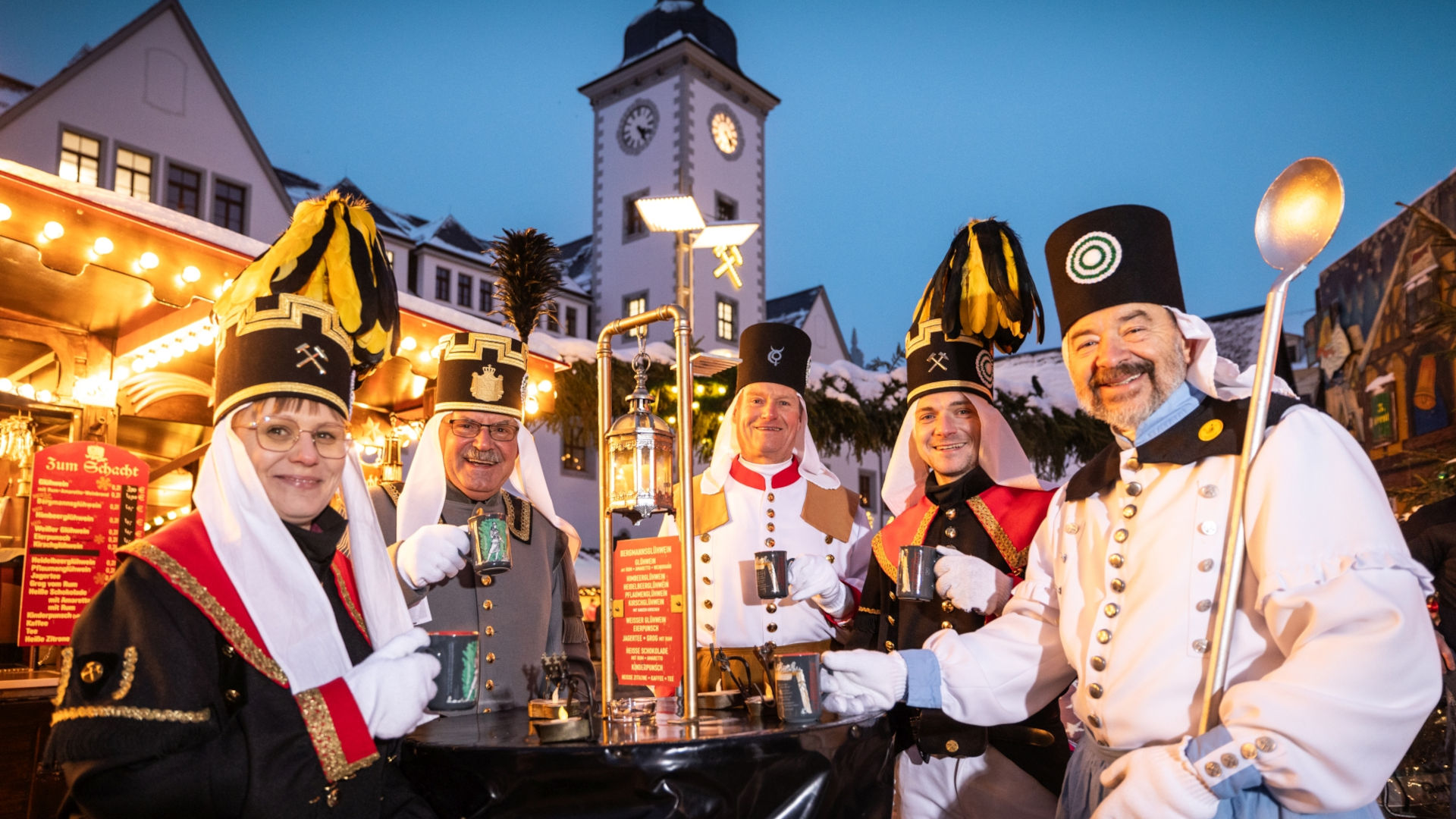 Vor stimmungsvoll beleuchteten Weihnachtsmarktständen stehen fünf Bergleute - vier Männer und eine Frau - im traditionell bergmännischen Habit mit Glühweintassen an einem Tisch. Es ist Abend. Im Hintergrund ist ein Kirchturm zu erkennen.
