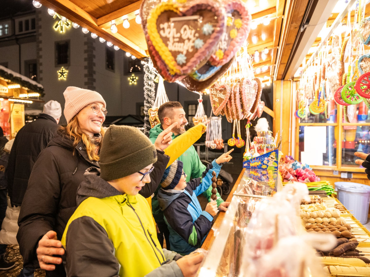 An einem beleuchteten Weihnachtsmarktstand mit verzierten Lebkuchenherzen und kandierten Früchten steht eine junge Frau mit ihrem Sohn und sieht sich lächelnd das Angebot an.