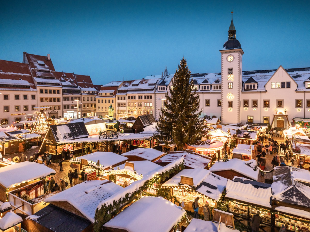 Blick von oben auf den abendlich erleuchteten Weihnachtsmarkt auf dem Freiberger Obermarkt mit schneebedeckten, beleuchteten Buden und einer großen Tanne in der Mitte