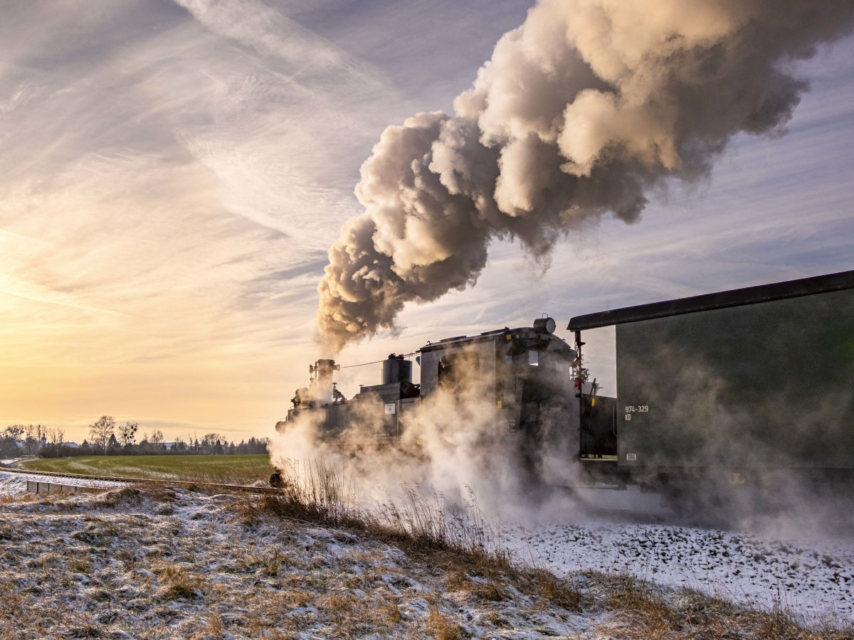 Ein historische Schmalspurbahn mit Dampflok fährt entlang einer Wiese, die mit winterlichem Raureif überzogen ist.