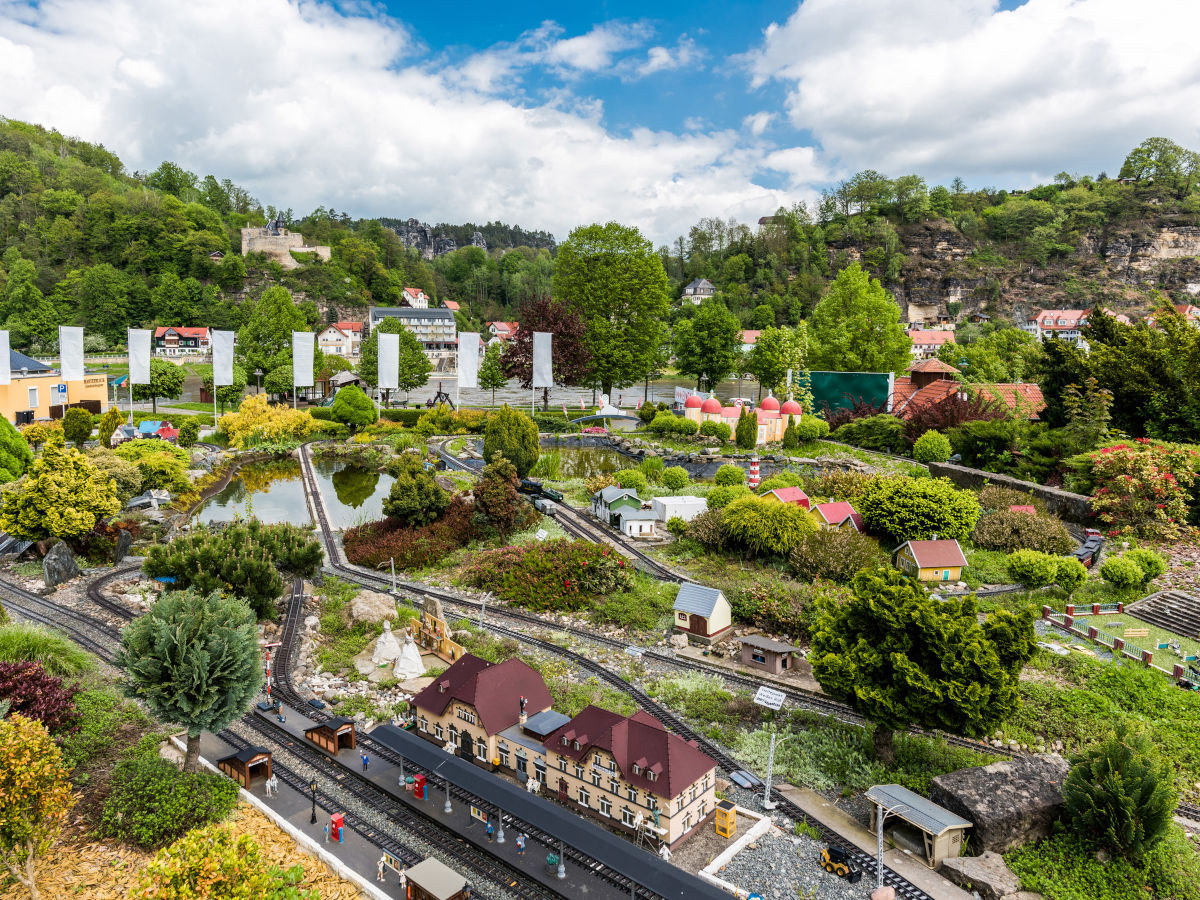 Vor den bizarren Felsen des Elbsandsteingebirges ist eine große Modellbahnanlage mit Schienen, Häusern und vielen grünen Büschen und Bäumen zu sehen.