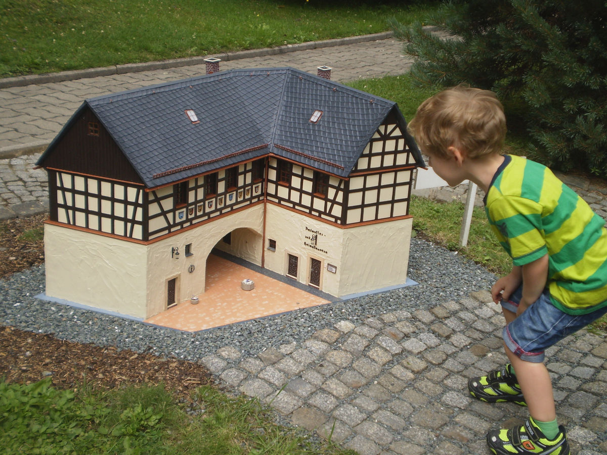 Ein kleiner Junge bückt sich, um sich das Miniaturmodell eines rechtwinkligen, cremefarbigen Fachwerkgebäudes mit einem Torbogen anzuschauen.