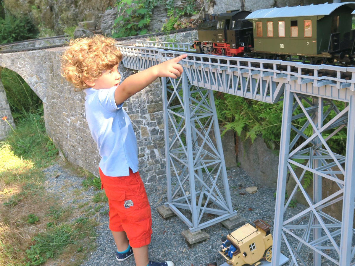 Ein kleiner Junge steht vor dem Modell einer Stahlbrücke. Diese ist genauso hoch wie der kleine Junge. Auf der Brücke fährt eine Modellbahn mit Lok und Wagen vorbei. Der kleine Junge zeigt begeistert darauf.