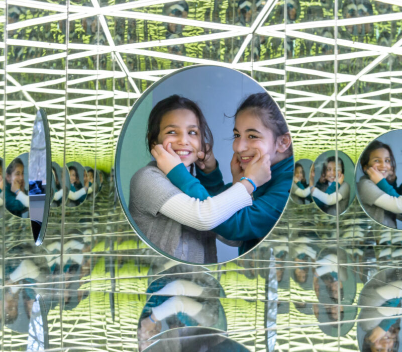 Zwei dunkelhaarige Mädchen von etwa zehn Jahren stehen nebeneinander hinter einem kreisförmigen Fenster und schauen in ein Spiegelkabinett, in dem sie sich unzählige Male spiegeln