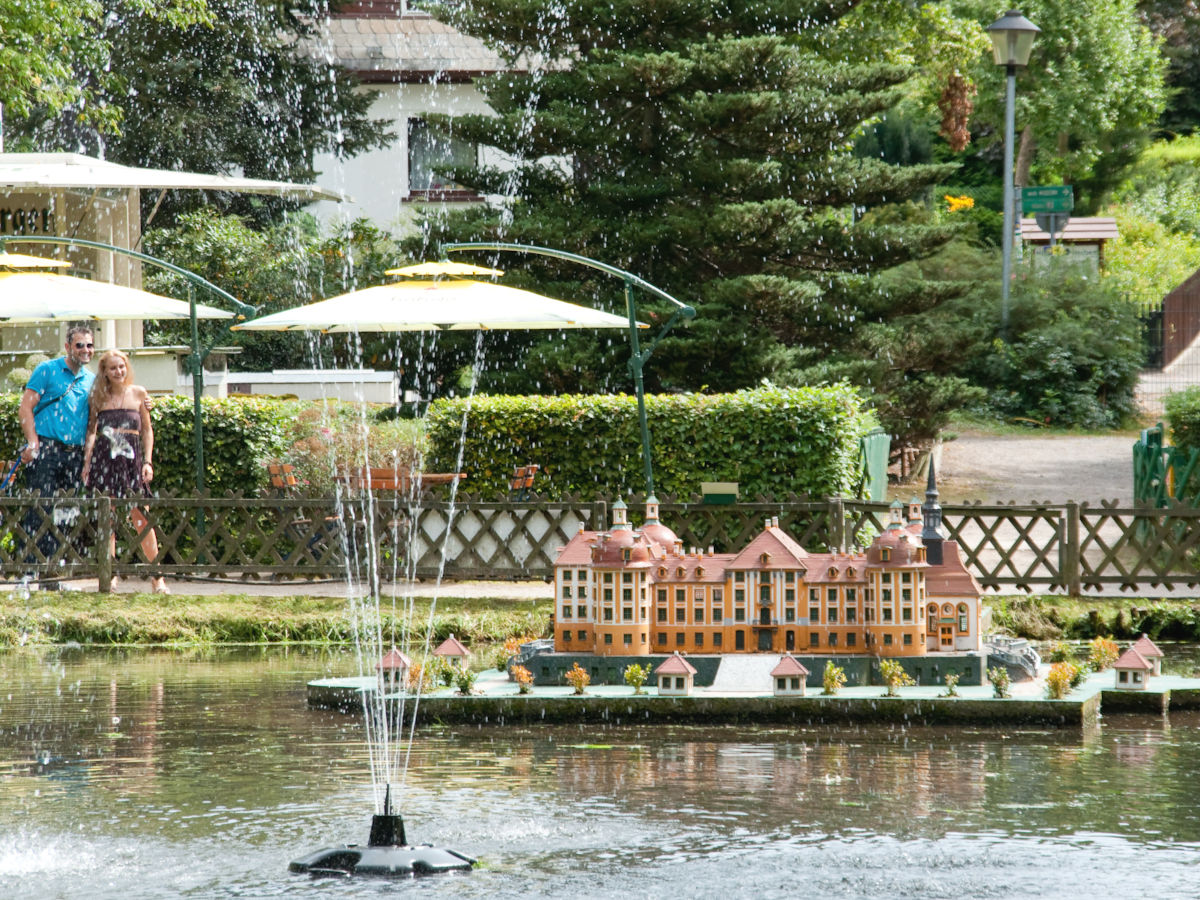 In einem Teich sprudelt eine Wasserfontäne. Dahinter befindet sich auf dem Teich eine Plattform, auf der ein Miniaturmodell des barocken sächsischen Schlosses Moritzburg mit seinen vier runden Türmen steht. 