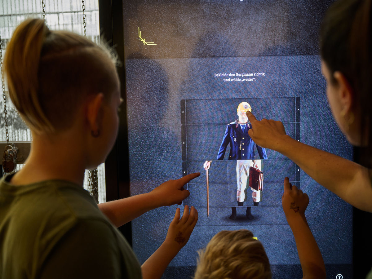 Zwei Kinder und ein Erwachsener stehen vor einem interaktiven Bildschirm mit dem Bild eines Bergmanns. Sie lösen eine Aufgabe, die auf dem Bildschirm angezeigt wird, und müssen den Bergmann mit den richtigen Kleidungsstücken ausstatten.