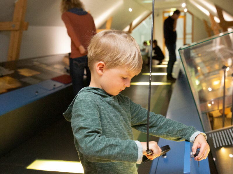 Ein kleiner blonder Lupe schaut in einem Museum durch eine Lupe auf ein Exponat.