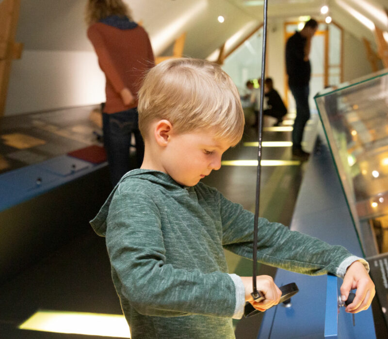 Ein kleiner blonder Lupe schaut in einem Museum durch eine Lupe auf ein Exponat.