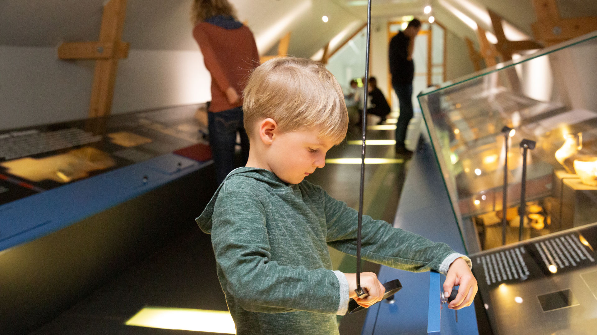 Ein kleiner blonder Lupe schaut in einem Museum durch eine Lupe auf ein Exponat.