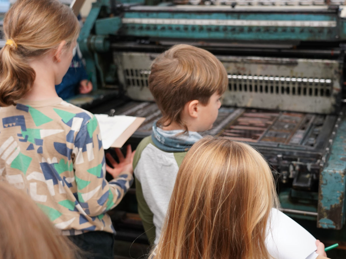 Drei Kinder mit Blöcken und Stiften in den Händen stehen vor einer historischen Druckmaschine.