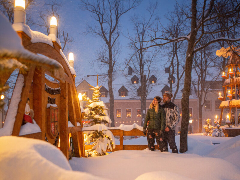 Eine Frau und ein Mann stehen in dicker Winterkleidung auf einem tiefverschneiten Marktplatz zwischen einem großen Schwibbogen aus Holz und einer großen Weihnachtspyramide aus Holz, die beide im Lichterglanz leuchten.