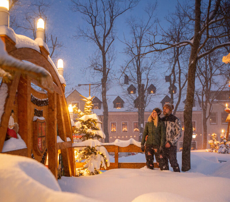 Eine Frau und ein Mann stehen in dicker Winterkleidung auf einem tiefverschneiten Marktplatz zwischen einem großen Schwibbogen aus Holz und einer großen Weihnachtspyramide aus Holz, die beide im Lichterglanz leuchten.