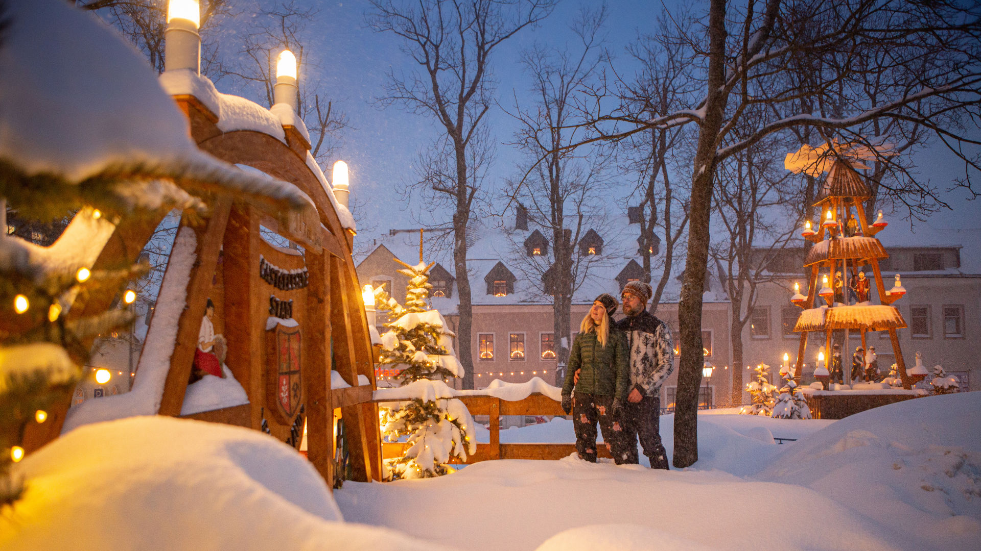 Eine Frau und ein Mann stehen in dicker Winterkleidung auf einem tiefverschneiten Marktplatz zwischen einem großen Schwibbogen aus Holz und einer großen Weihnachtspyramide aus Holz, die beide im Lichterglanz leuchten.