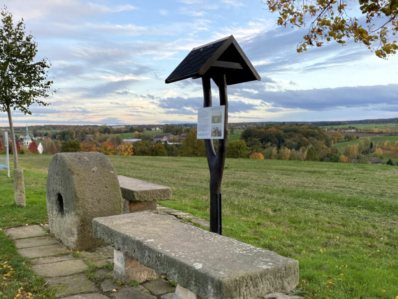 zwei Steinbänke stehen neben einem alten Mühlstein und einem Wegweiser aus Holz an einem Wanderweg oberhalb einer Wiese mit weitem Panoramablick