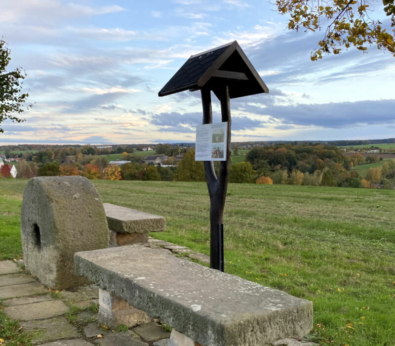 zwei Steinbänke stehen neben einem alten Mühlstein und einem Wegweiser aus Holz an einem Wanderweg oberhalb einer Wiese mit weitem Panoramablick
