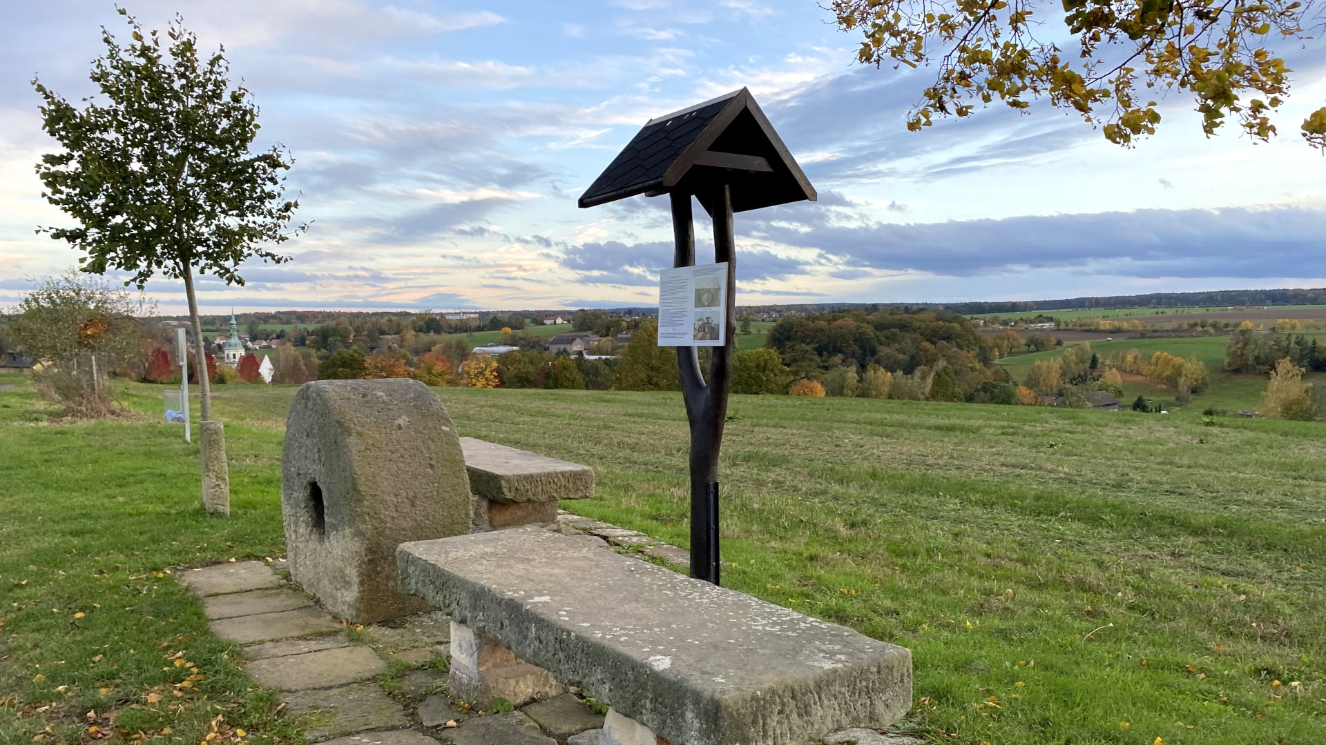 zwei Steinbänke stehen neben einem alten Mühlstein und einem Wegweiser aus Holz an einem Wanderweg oberhalb einer Wiese mit weitem Panoramablick