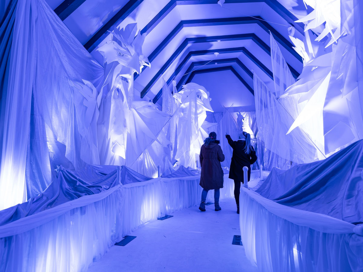 Ein mit weißen Tüchern verkleideter Tunnel wird mit blau-lila Licht angeleuchtet, sodass der Eindruck eines Eistunnels entsteht. Zwei Personen gehen hindurch.