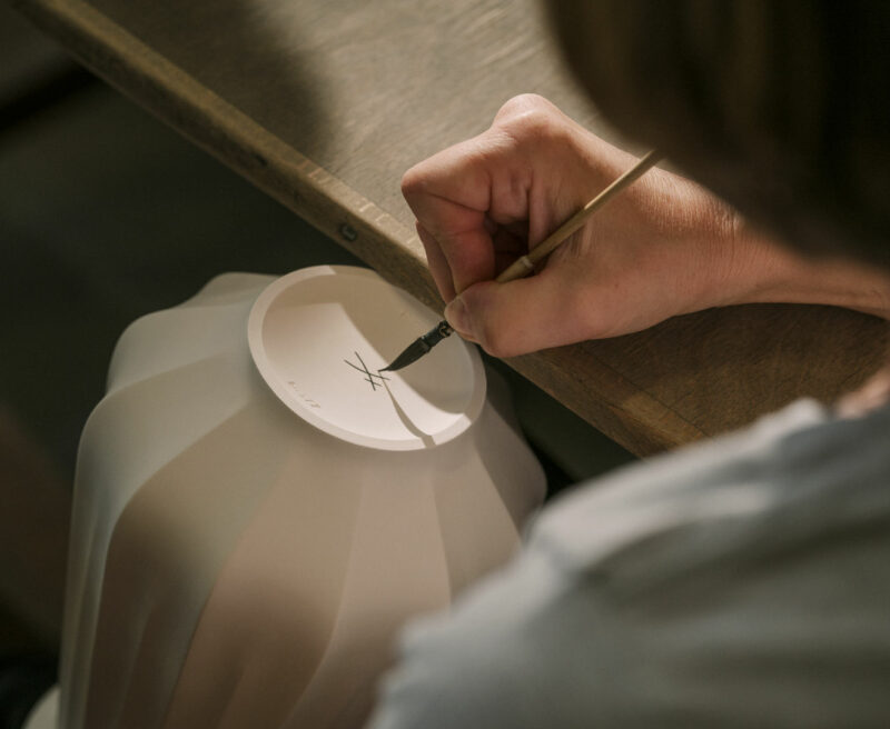 Eine Hand einer nur im Anschnitt von hinten zu sehenden Frau hält einen feinen Pinsel, mit dem auf den Boden einer weißen, unglasierten Vase die gekreuzten Schwerter, das Markenzeichen des Meissener Porzellans, aufgemalt werden.