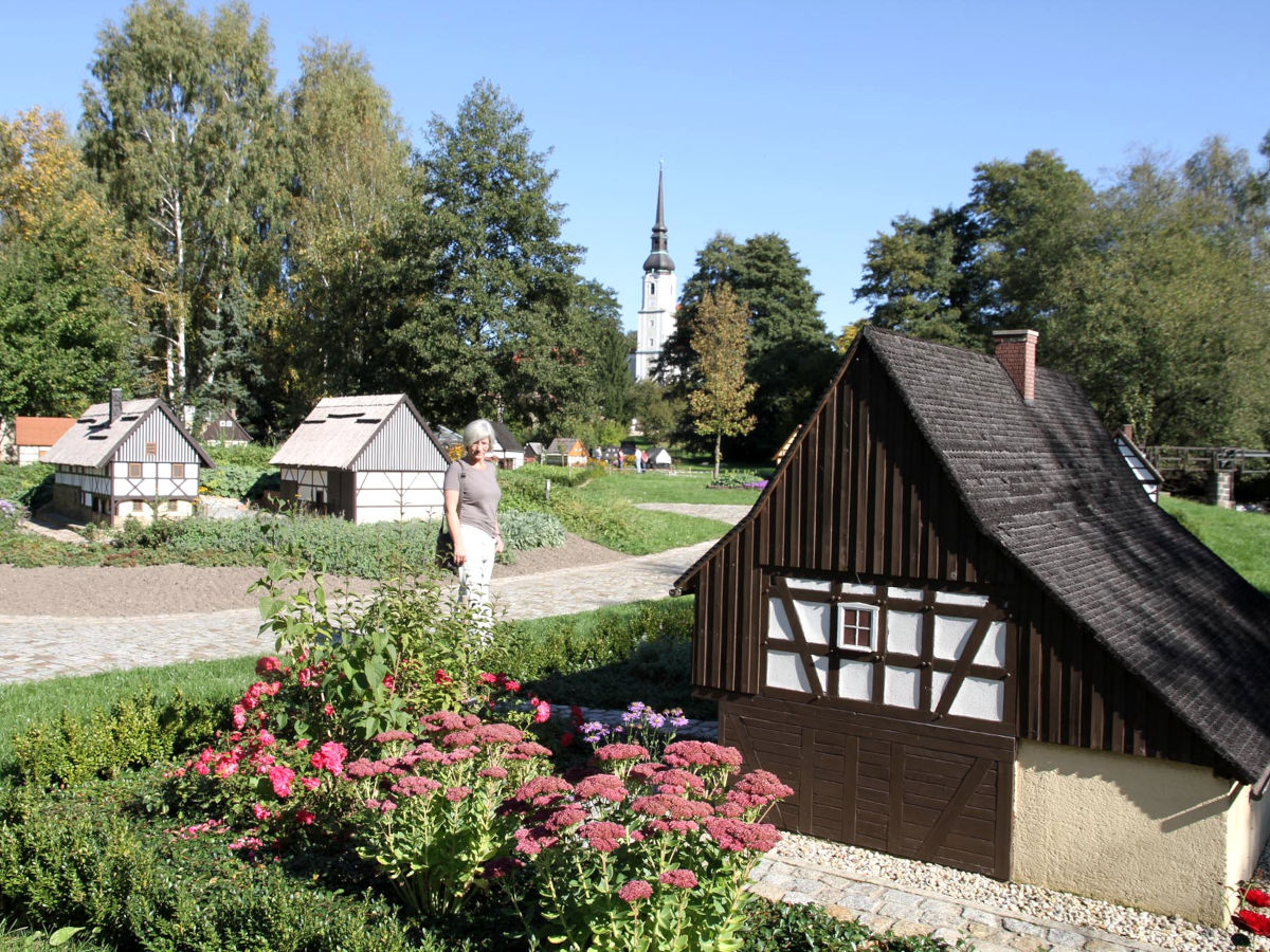 In einem Park mit einem Kirchturm im Hintergrund steht ein mit dunklem Holz verkleidetes historisches Fachwerkhaus im Miniaturformat. Eine Frau betrachtet es.